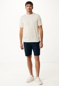 Hellebeige Kurzarm-T-Shirt mit Rundhalsausschnitt, kombiniert mit marineblauen Shorts und weißen Turnschuhen, vor einem schlichten weißen Hintergrund.