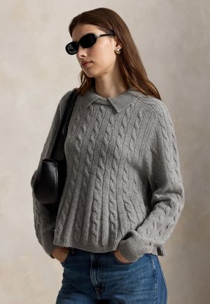Femme portant des lunettes de soleil noires, un pull gris torsadé avec col, un jean bleu, des boucles d’oreilles dorées et un sac à bandoulière noir.