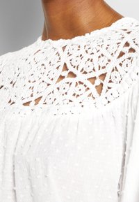 Hvid bluse med et tekstureret stof med subtile prikker, en blonde yoke med indviklede blomstermønstre og en afslappet pasform.