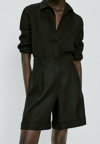 Personne portant un short noir taille haute avec revers et une chemise noire à manches longues boutonnée avec les manches retroussées.