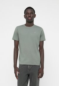 Tricou verde din bumbac, cu guler rotund și mâneci scurte. Prezintă un mic logo pe piept. Asortat cu blugi din denim gri.