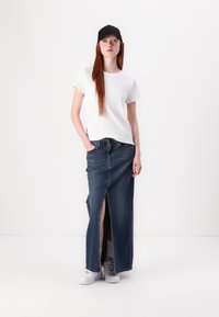 Maglietta bianca, gonna maxi in denim a vita alta con spacco frontale e sneakers bianche. La gonna è realizzata in un classico denim blu. Cappellino nero.