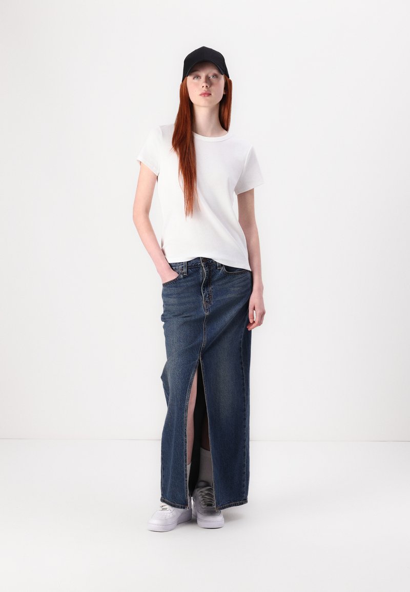 Weißes T-Shirt, hochgeschnittener Denim-Maxirock mit Frontschlitz und weißen Sneakers. Der Rock besteht aus klassischem blauen Denim-Material. Schwarze Kappe.