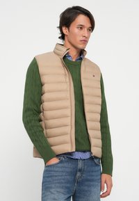Beige padded vest over a green cable-knit sweater, with a blue checkered shirt underneath. Features a high collar and side zipper.