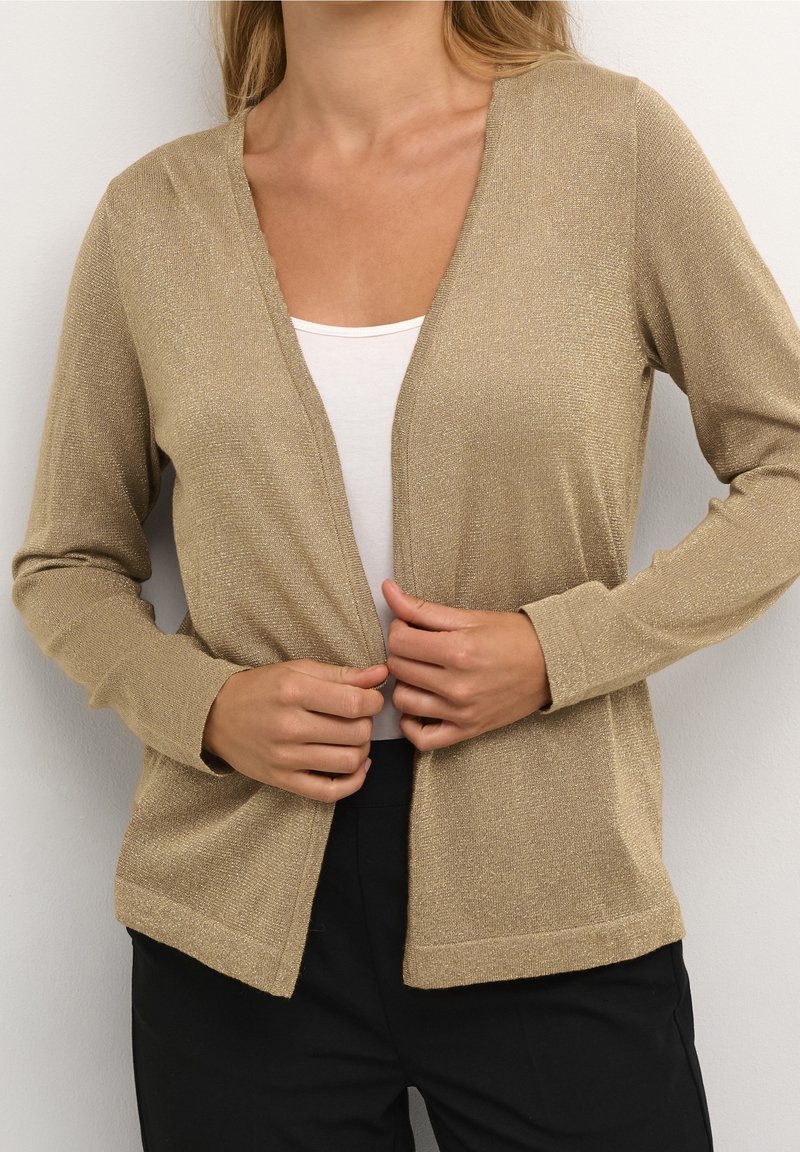 Femme portant un cardigan beige scintillant ouvert sur un haut blanc et un pantalon noir, ajustant le cardigan avec les deux mains.