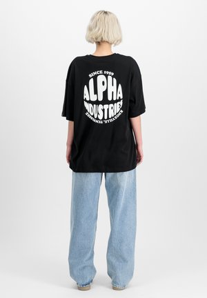 T-shirt noir oversize avec un texte circulaire blanc "ALPHA INDUSTRIES" et un jeans baggy bleu clair. Design simple et décontracté.