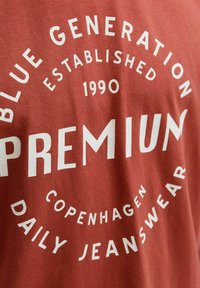 Rudai spalvos medžiagos marškinėliai su baltais užrašais apvaliu dizainu, kuriuose rašoma "BLUE GENERATION", "PREMIUM" ir "COPENHAGEN DAILY JEANSWEAR."