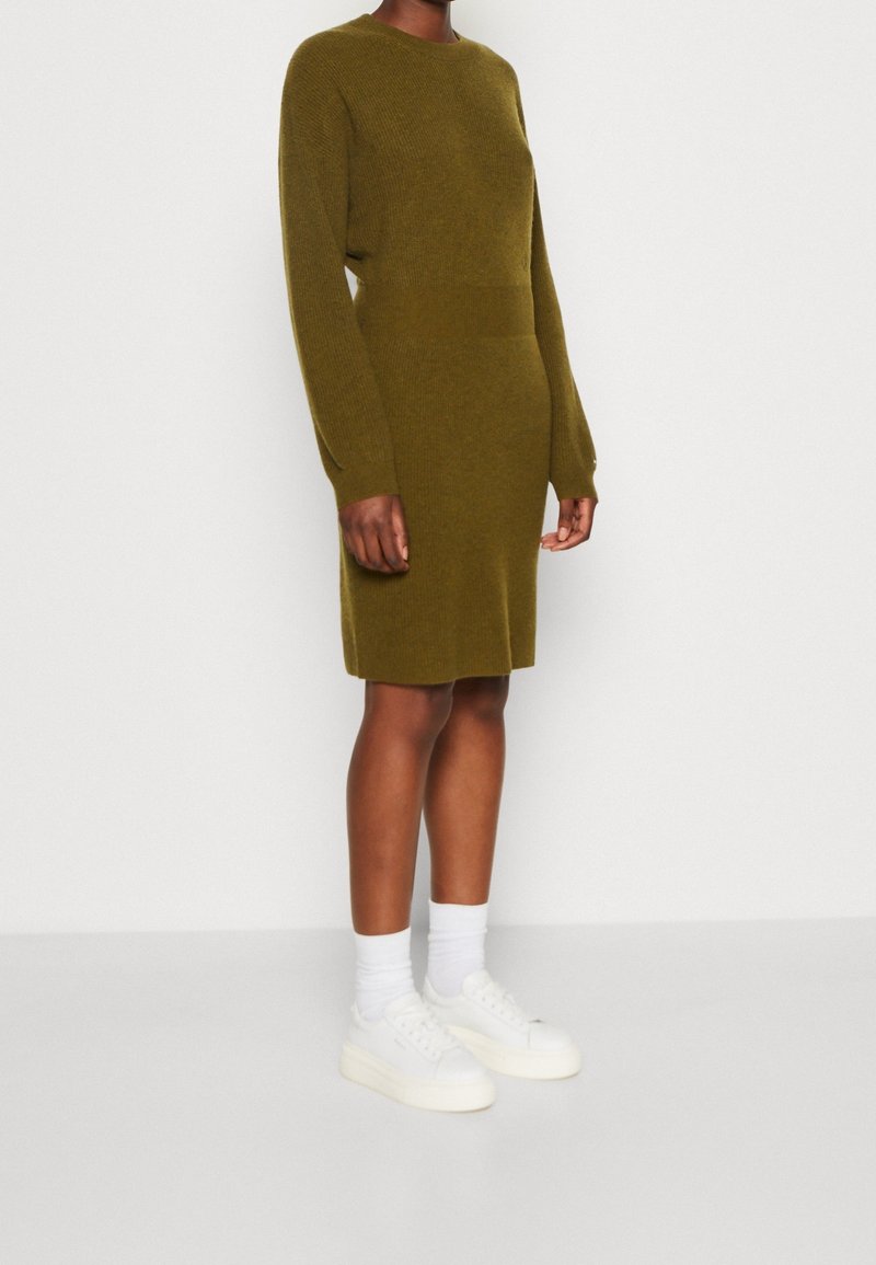 Personne à la peau foncée portant une robe en maille vert olive mi-longue, des chaussettes blanches et des baskets blanches, debout devant un fond clair uni.