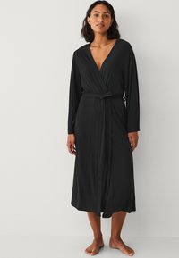 Robe noire enroulée avec des manches longues, décolleté en V profond et taille ceinturée. Fabriquée dans un tissu doux et fluide, elle arrive au mollet et présente une texture lisse.