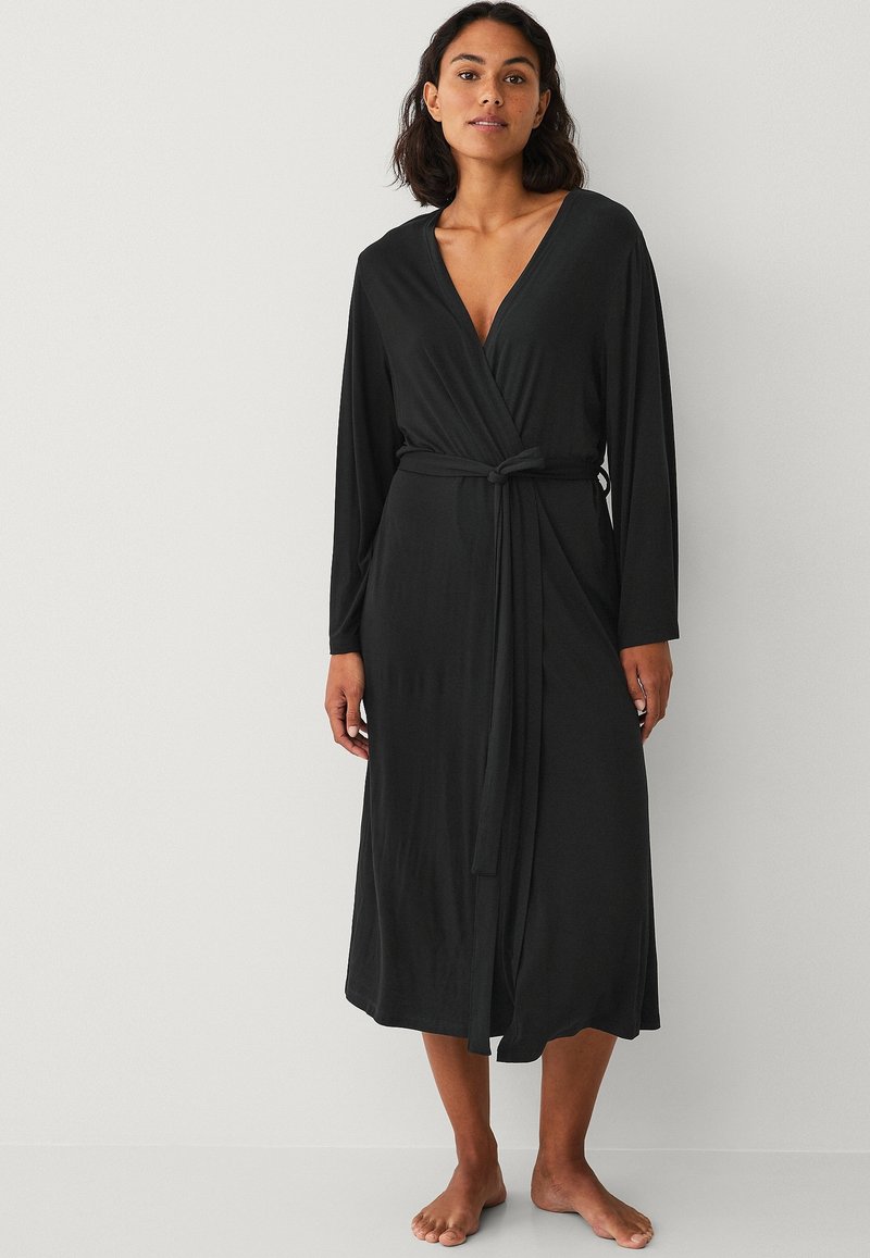 Robe noire enroulée avec des manches longues, décolleté en V profond et taille ceinturée. Fabriquée dans un tissu doux et fluide, elle arrive au mollet et présente une texture lisse.