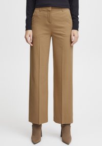 Beige weit geschnittene Hose mit glatter Textur, flachem Bund, Gürtelschlaufen und zwei Seitentaschen, kombiniert mit beigefarbenen spitz zulaufenden Ankle Boots.