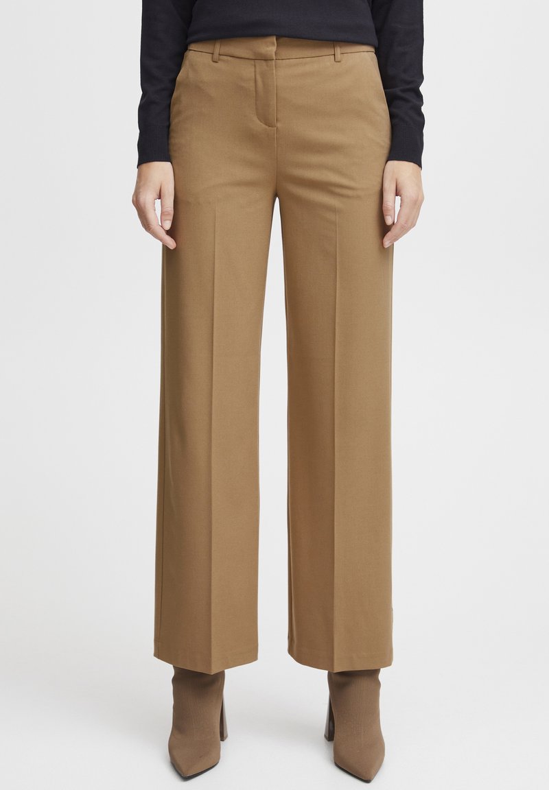 Beige weit geschnittene Hose mit glatter Textur, flachem Bund, Gürtelschlaufen und zwei Seitentaschen, kombiniert mit beigefarbenen spitz zulaufenden Ankle Boots.