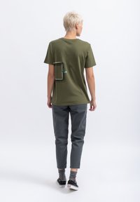 T-shirt vert olive à manches courtes, avec un design de poche arrière rectangulaire en noir et en bleu canard. Associe à un pantalon gris fuselé et des baskets noires.