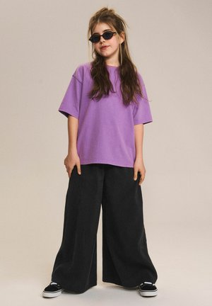 Młoda dziewczyna z długimi włosami, nosząca czarne okulary przeciwsłoneczne, luźny fioletowy t-shirt, szerokie czarne spodnie i czarne trampki, stojąca na jednolitym tle.