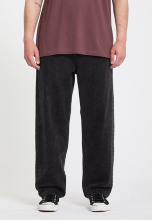 Personne portant un jean noir ample, des baskets noires et blanches, et une chemise bordeaux, debout devant un fond blanc.