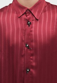 Maroon striped satin shirt with decorative black beaded flower-shaped buttons fastened at the collar and front.