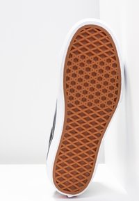 Svart skate-sko med en vit gummimellansula och en texturerad brun gummiyttersula. Har ett geometriskt greppmönster för gott fäste.