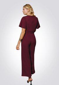 Bordový overal s krátkými volánkovými rukávy, vypasovaným pasem a širokými nohavicemi, vyrobený z hladkého materiálu. Zadní část má diskrétní zapínání.