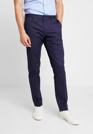 Man draagt donkerblauwe slim fit nette broek, lichtblauw overhemd en witte casual sneakers, staand tegen een egale lichte achtergrond.