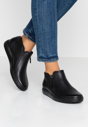 Ankelhøje sorte lædersko med lynlås båret med blå, opsmøgede, tætsiddende jeans på en glat, hvid gulv.