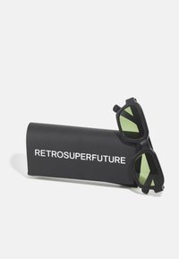 Črne sončne očala z zelenimi lečami in geometrijskim dizajnom, položena na črno etui, na katerem je v belih črkah napisan zapis "RETROSUPERFUTURE".