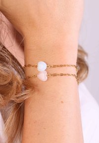 Gouden armband met een ketting featuring twee hartvormige edelstenen, één blauwe en één roze, met een gladde textuur en een delicaat ontwerp.
