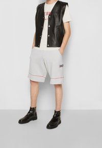 Gilet in pelle nera senza maniche sopra una t-shirt color crema, abbinato a pantaloni corti grigi con dettagli rossi e stivali neri con zip alla caviglia.