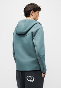 Νεαρός άνδρας με κοντά σγουρά μαλλιά που φοράει πράσινη φούτερ με κουκούλα και μαύρο παντελόνι με λευκό λογότυπο Nike στην τσέπη, στέκεται με τα χέρια στις τσέπες, κοιτάζοντας προς τα δεξιά.