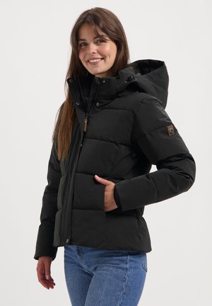 Young woman smiling, wearing a black hooded puffer jacket with zipper and snap buttons, and blue jeans, standing against a plain background.