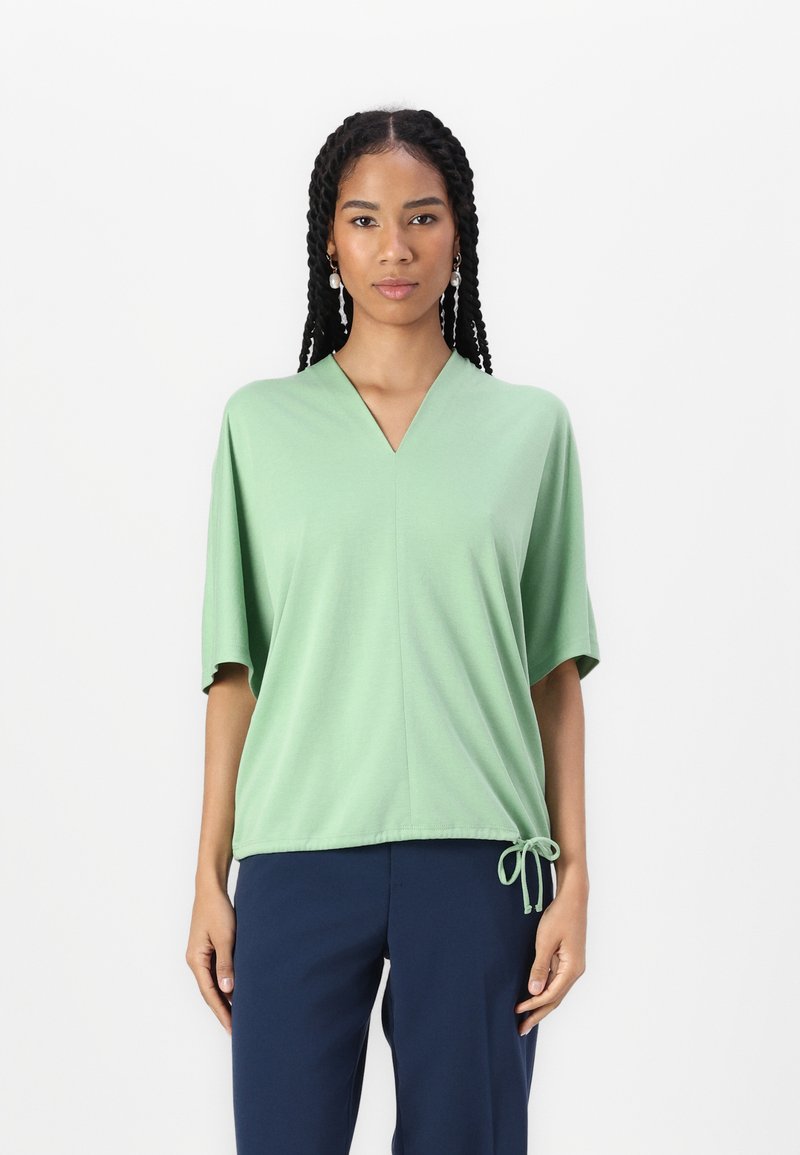 Top verde chiaro a maniche corte con scollo a V, caratterizzato da un cordoncino sull'orlo. Tessuto morbido con una consistenza liscia. Indossato con pantaloni blu navy.