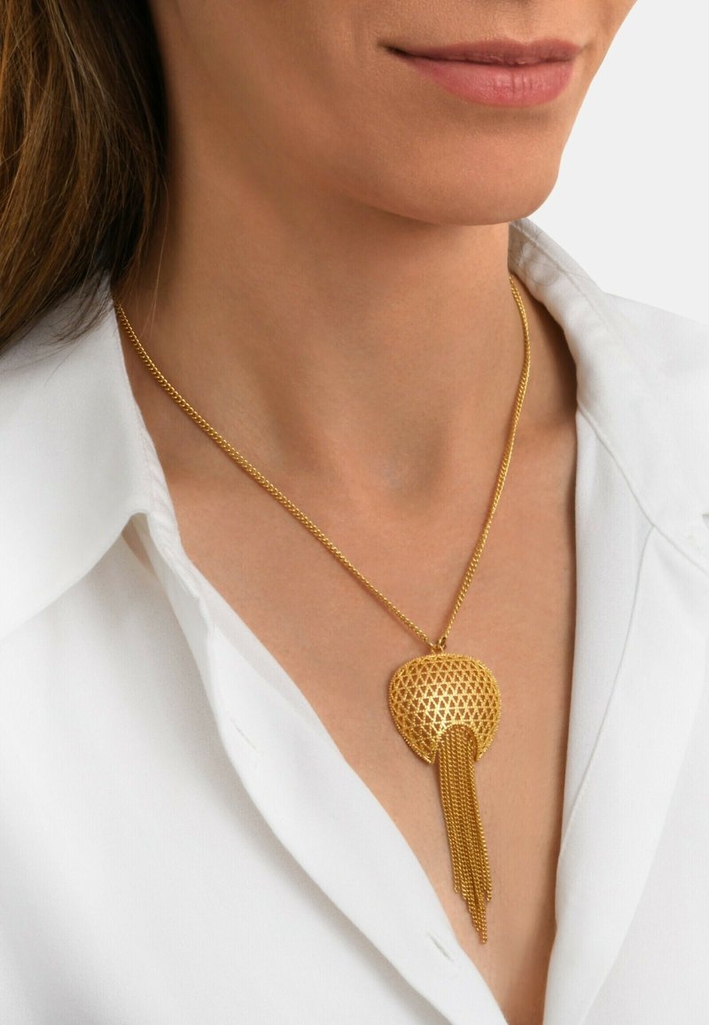 Collier doré avec un pendentif texturé à motif géométrique et des franges allongées, porté sur une chemise blanche à col.