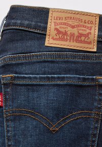 Un bolsillo trasero de unos vaqueros de mezclilla azul oscuro con costuras naranjas y una etiqueta de cuero color marrón de Levi Strauss & Co. que muestra dos caballos y texto.