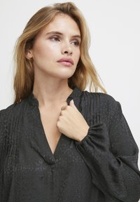 Blusa nera con scollatura a V, dotata di pieghe e maniche a sbuffo. Il materiale presenta una texture e un motivo sottili, offrendo un aspetto liscio.