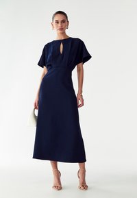 Tummansininen, midi-pituinen mekko lyhyillä liehuvilla hihoilla ja avokaula-aukolla. Tehty sileästä kankaasta, yhdistettynä nude-korkokenkiin ja valkoiseen käsilaukkuun.