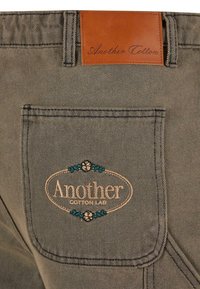 Grijze katoenen broek met een leren label met de tekst "Another Cotton." Voorzien van een geborduurd logo met bloemenaccenten op de achterzak.