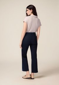 Blusa corta a maniche corte color beige chiaro e pantaloni svasati di un blu scuro. I pantaloni presentano una texture liscia e tasche posteriori. Il modello indossa scarpe beige.
