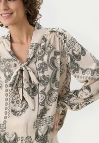 Beige blus med svart paisleymönster, knytkrage och långa ärmar. Slät väv med rynkade axlar och lös passform.