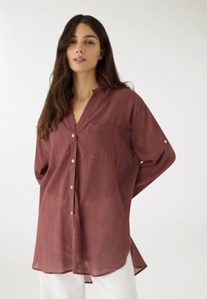 Donna che indossa una camicia ampia a maniche lunghe, abbottonata, di colore mauve scuro con trama, abbinata a pantaloni bianchi su sfondo bianco.