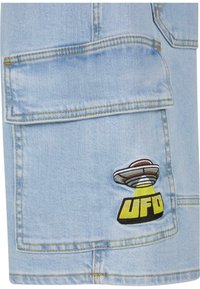 Lyseblå denimstoff har et stort broderi av et romskip i grått, brunt og gult, med ordet "UFO" i fet skrift.