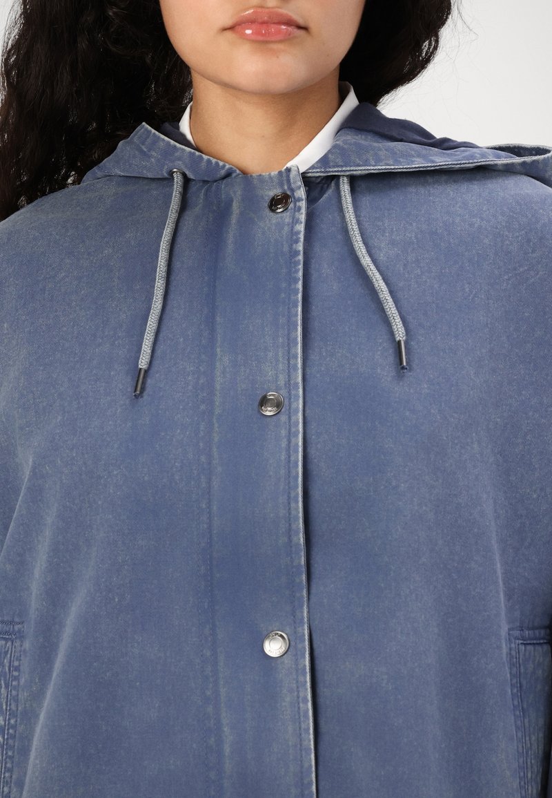 Veste en denim bleu clair avec capuche, dotée d'une fermeture à pression à l'avant et de cordons réglables au col pour un ajustement personnalisé.