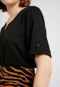 Kvinde iført sort kortærmet bluse med optrillede ærmer og knapdetalje, parret med en brun og sort tigermønsteret nederdel og guldhalskæde.