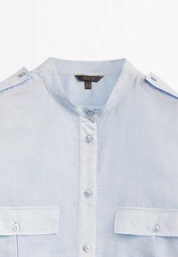 Chemise bleu clair à col, avec boutons devant, deux poches poitrine à rabats et épaulettes boutonnées, présentée sur fond blanc.