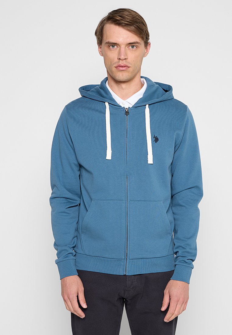 Młody mężczyzna ubrany w niebieską bluzę na zamek z białymi sznurkami, noszący białą koszulę, stoi na jasnym tle.