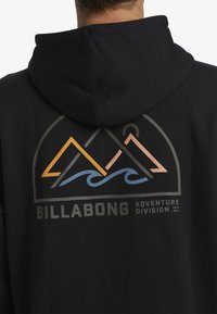 Černá mikina s kapucí s grafickým designem zobrazujícím hory, vlny a text "BILLABONG ADVENTURE DIVISION" v tlumených barvách.