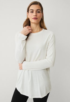 Ung kvinde med langt brunt hår iført gyldne hoop øreringe og en hvid langærmet top med rund hals foran en ensfarvet baggrund.