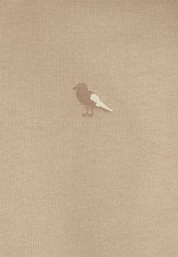 Beige knit fabric featuring a small bird emblem in light brown and white at the center. Smooth texture with a simple, subtle design.