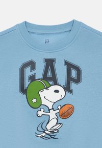 T-shirt de mangas curtas azul claro com um design gráfico de um cão em cartoon com um capacete verde, segurando uma bola de futebol laranja com o texto "GAP" acima.