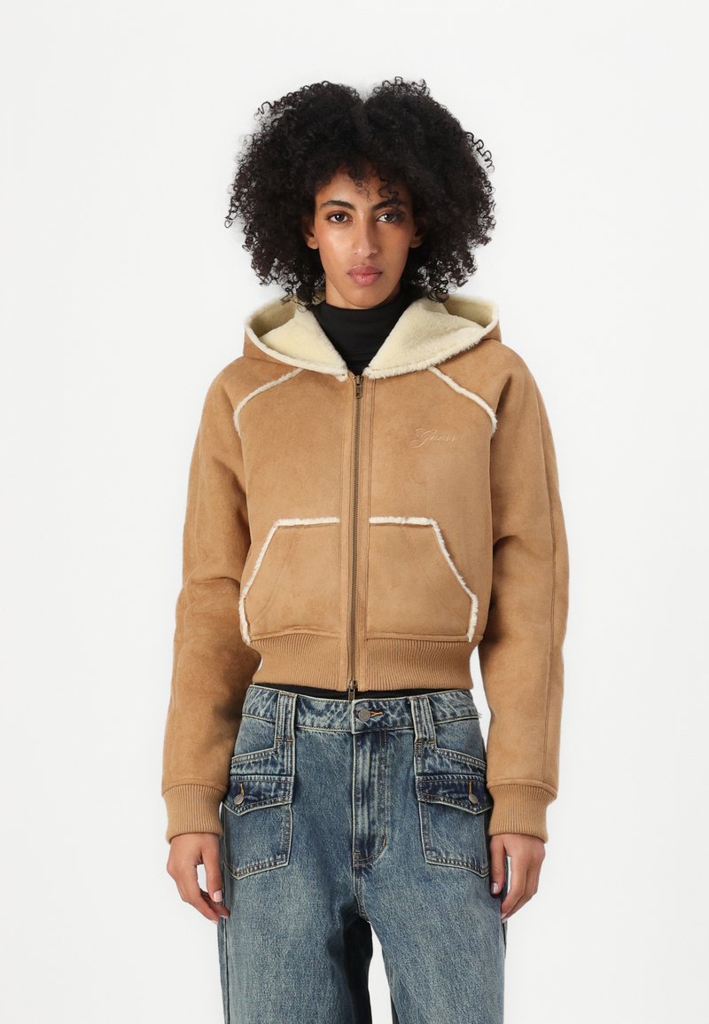 Veste bombardier en daim marron avec col en fausse fourrure, fermeture éclair à l'avant et deux poches avant, présentant des coutures contrastantes blanches et des poignets côtelés.
