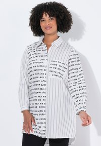 Donna con capelli ricci che indossa una camicia lunga bianca con strisce verticali nere e motivi testuali, mentre sorride con una mano leggermente sollevata.