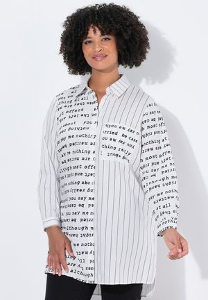 Femme aux cheveux bouclés portant une longue chemise blanche à rayures verticales noires et motifs textuels, souriante avec une main légèrement levée.